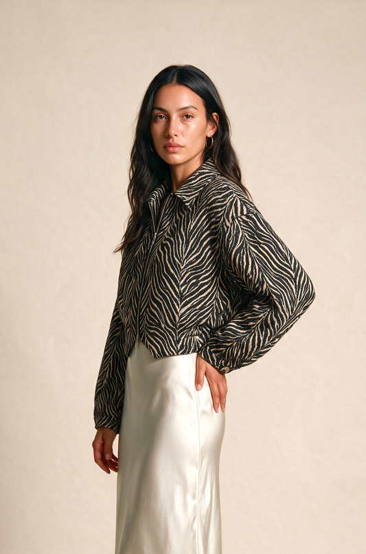 A woman stands in a studio wearing a black and white zebra-print long-sleeve blouse and a shiny cream satin maxi skirt, paired with light-colored flat sandals.