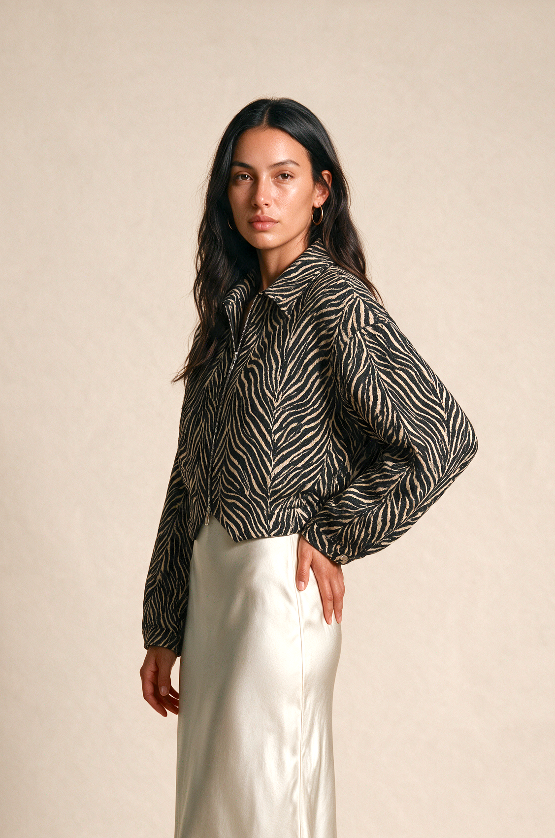 A woman stands in a studio wearing a black and white zebra-print long-sleeve blouse and a shiny cream satin maxi skirt, paired with light-colored flat sandals.