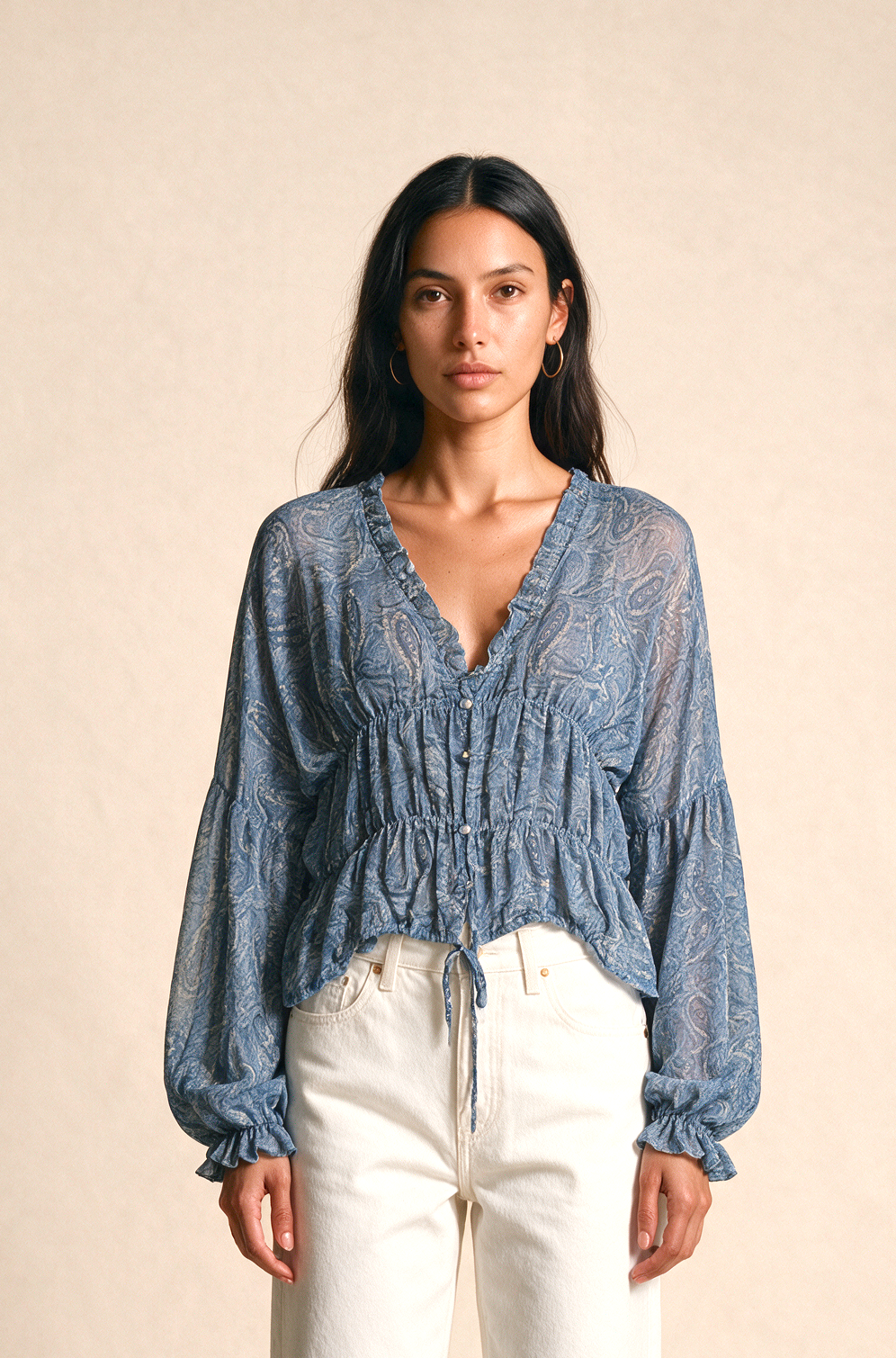 A woman stands against a warm beige studio backdrop wearing a blue smocked long-sleeve blouse with white wide-leg pants and flat sandals.