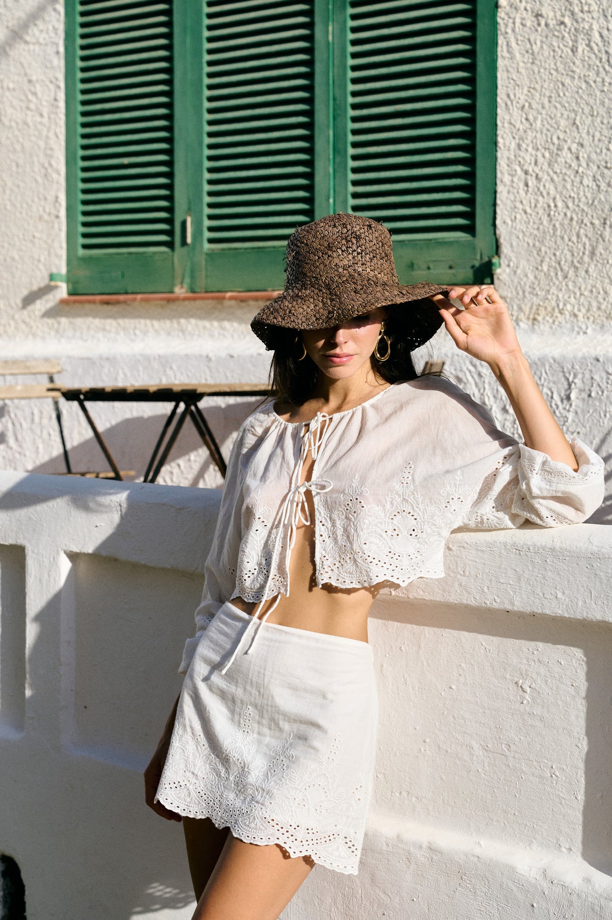 A woman wearing a white eyelet two-piece set with a tie-front crop top and matching mini skirt, styled with a brown woven sun hat, leaning against a white stucco wall in front of green shutters.