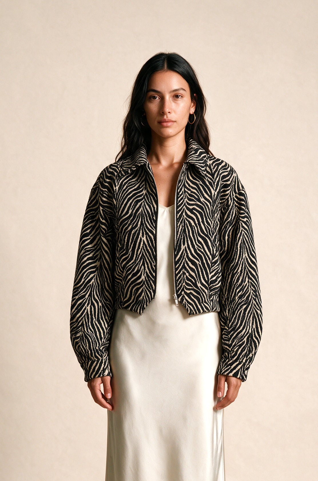 A woman stands against a plain beige studio background wearing a black-and-white zebra-print cropped jacket over a white satin midi skirt, with neutral sandals.