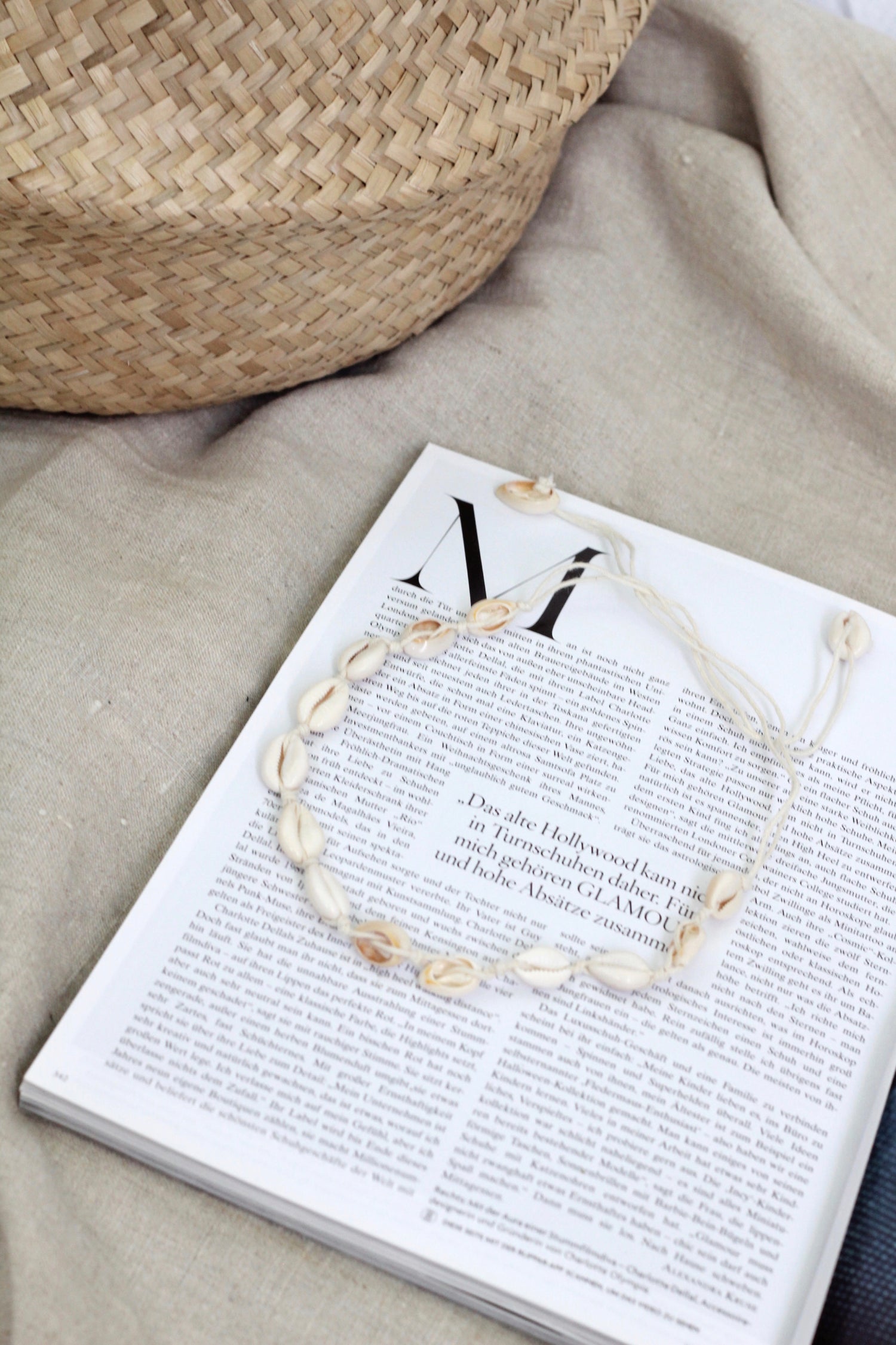 A beige-colored shell choker with real shells, displayed on a textured surface next to a straw hat and a fashion magazine.